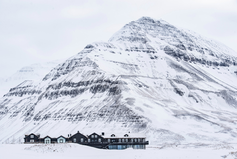 A large dark lodge sits at the base of a snow covered mountain. The lodge itself is snow covered wit